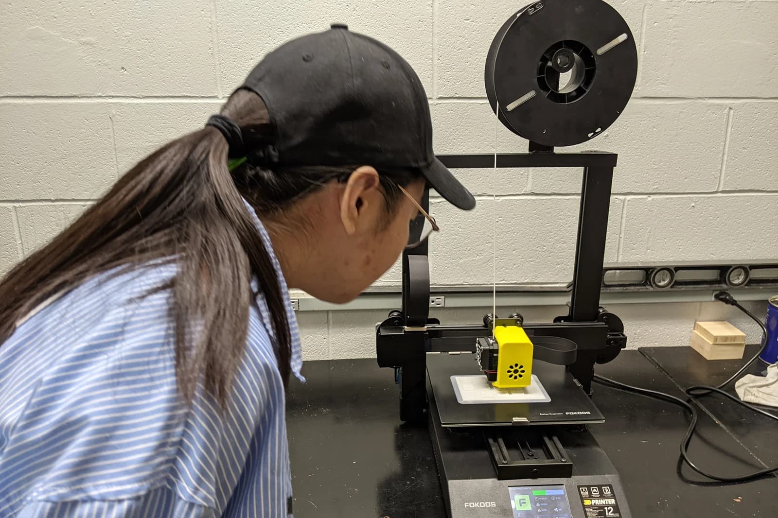 Xinxin Li leans toward a desktop filament printer to observe a white object being printed on its build plate.