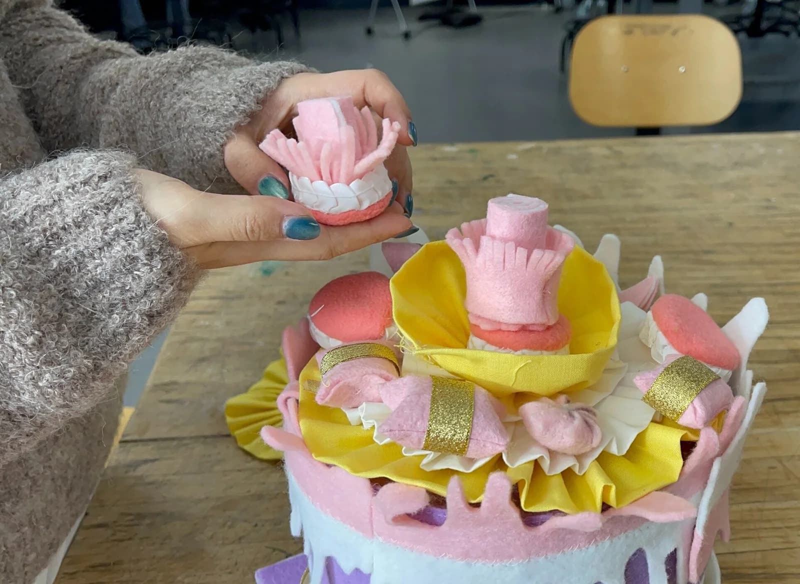 Hands arranging fabric dessert pieces on a layered soft cake at a worktable.