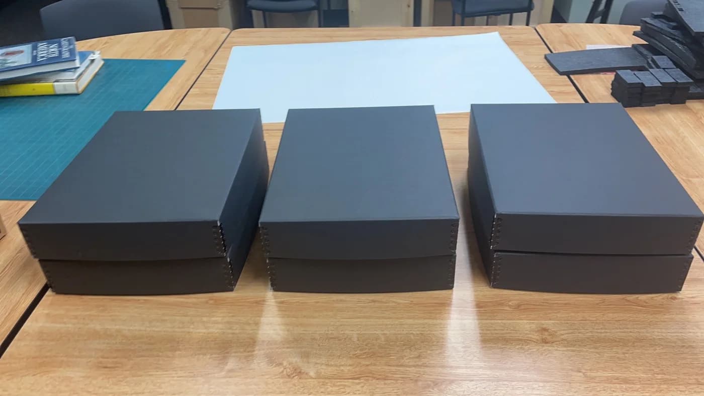 Three closed grey rectangular boxes are lined up on a workshop table.