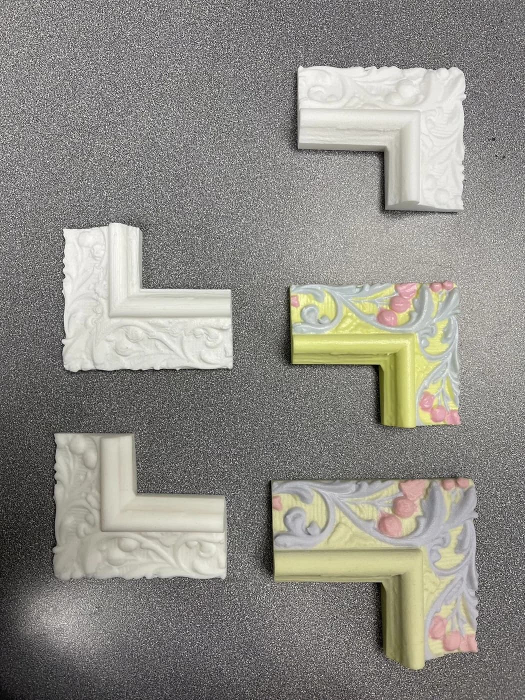 Four ornate frame-corner pieces, two white and two multicoloured, are arranged on a grey surface.
