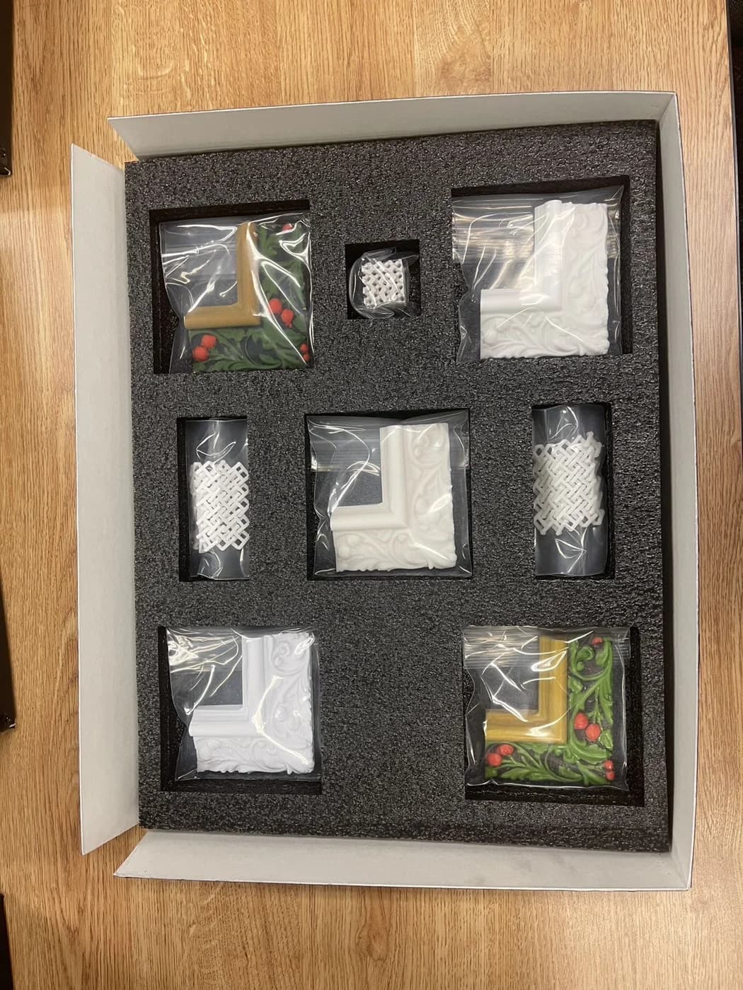 Bagged printed objects, including colourful frame corners, sit in individual cutouts within a grey presentation box.