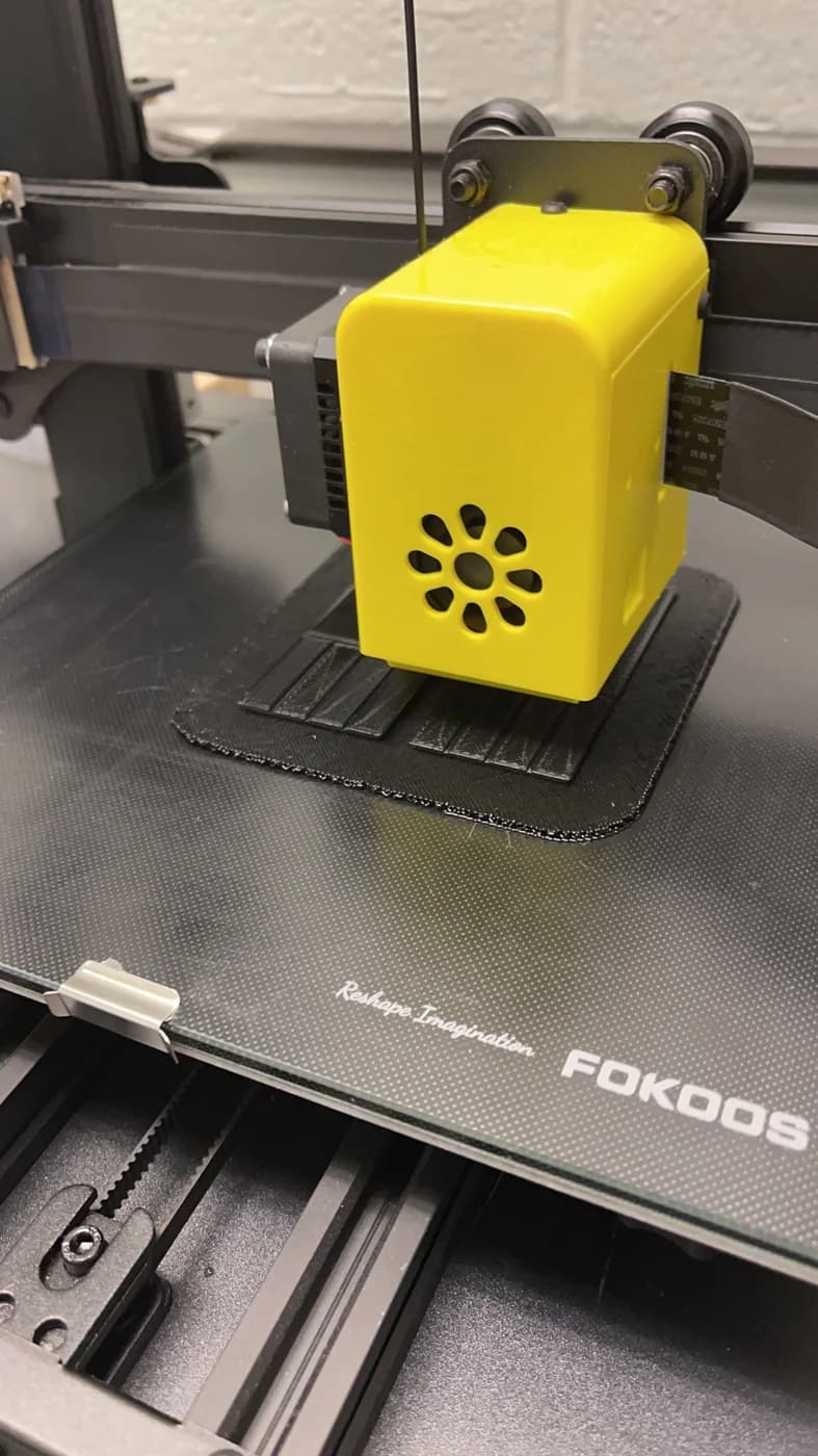 A yellow printer head sits above a dark L-shaped frame-corner print on a build plate.