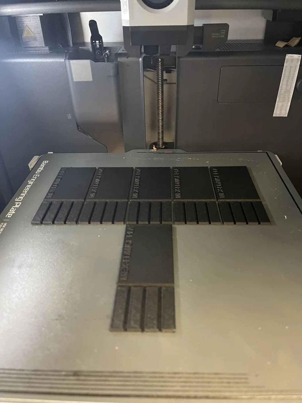Rows of dark rectangular pieces form a T-shaped arrangement on a printer build plate.