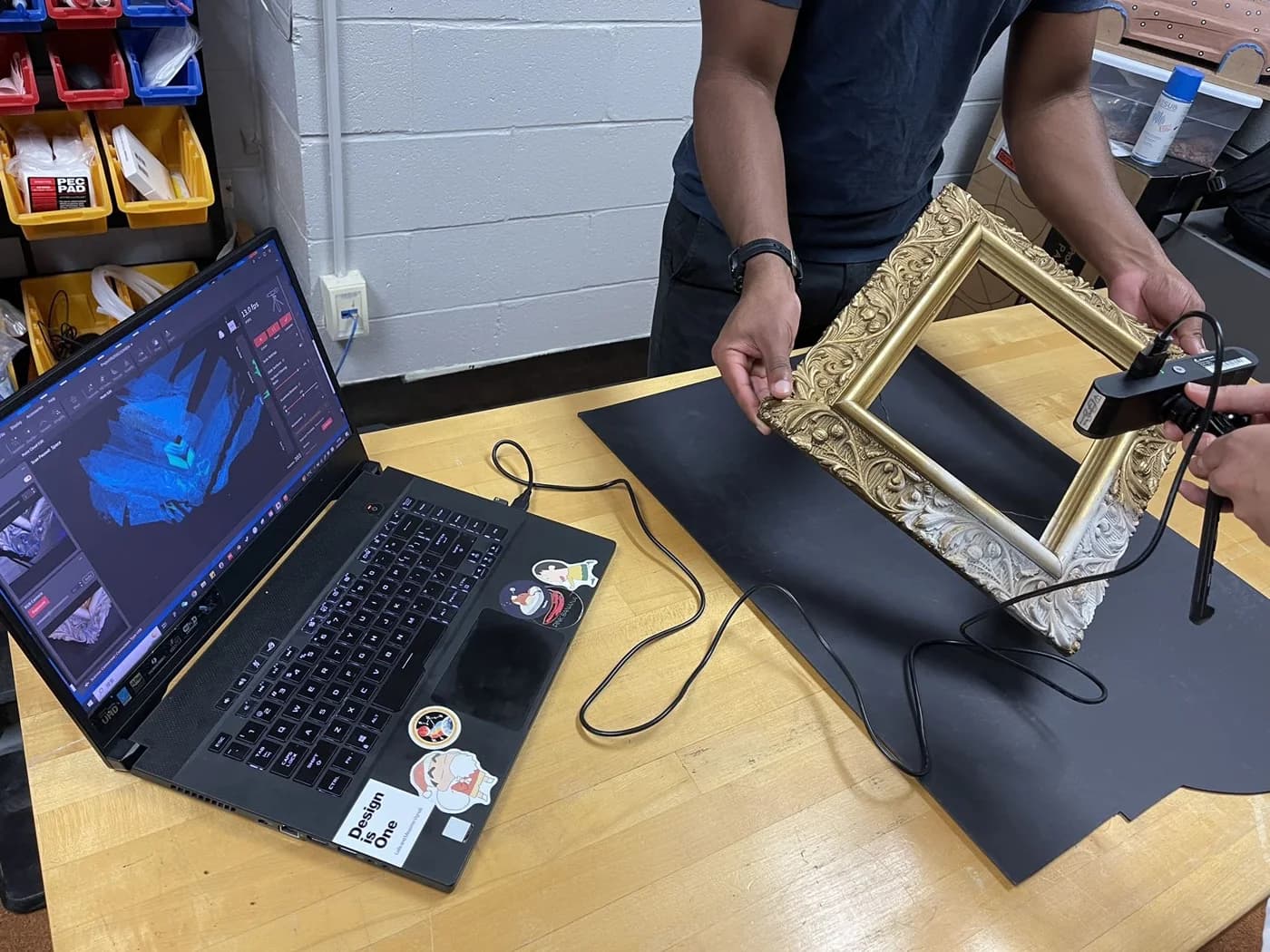 A handheld scanner is aimed at an ornate frame beside a laptop displaying blue scanned geometry.
